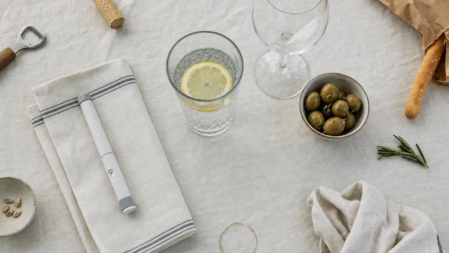 Overhead view of sparkling water with lemon beside an empty wine glass, olives and a capped injection pen on linen.