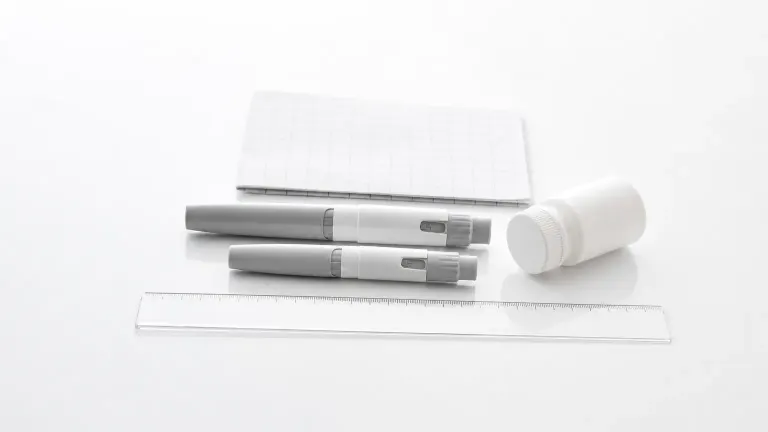 Two injection pens, a pill bottle and an unmarked ruler on a white surface