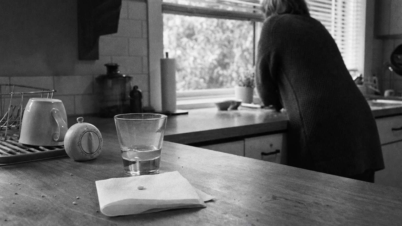 Black-and-white photo of a single tablet, a small glass of water and a kitchen timer at dawn.